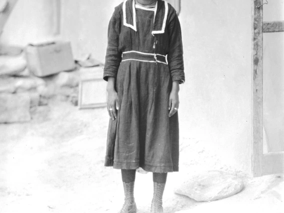 a young Hopi woman with short hair is standing outside a house and looking into the camera. She is wearing a dark, long-sleeved dress. Her button-up boots go up to her calves.