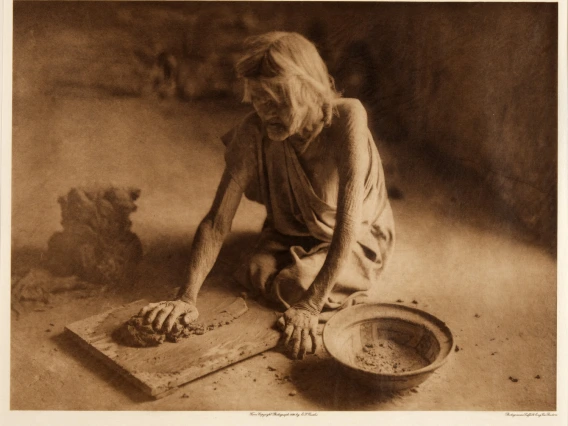 an elderly Hopi woman kneels on the ground and is kneading clay with her hands. To her side is a bows with an everted rim.