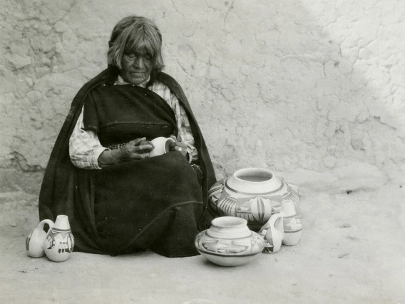 a Hopi woman sits on the ground in front of an adobe wall, sanding a small ceramic vessel she is sanding. She is surrounded by other vessels she has already made.