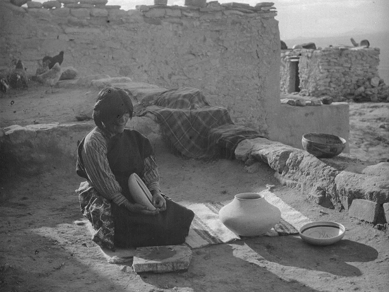 A Hopi woman sits on a woven textile which is spread on the ground. She is holding and is surrounded by pieces of pottery she has made.