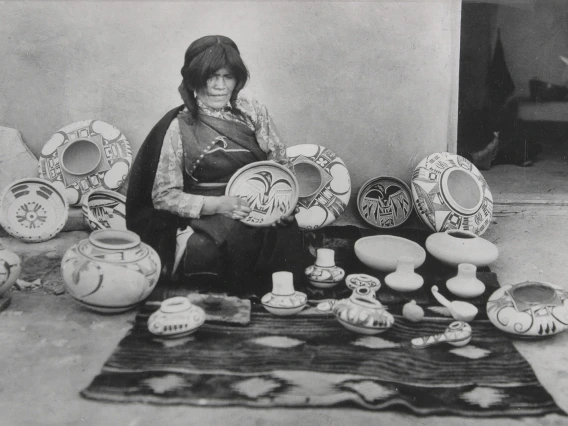 a Hopi woman sits on the ground with a blanket spread before her and a great number of pots she has made surround her