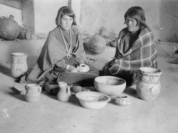 a young Hopi woman with short hair shits on the ground looking at pottery she and her mother, to her left, have made together