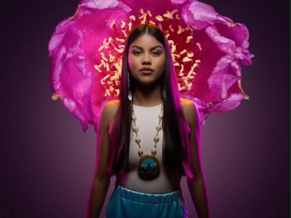 a young woman with long dark hair stands in front of a large bright pink flower