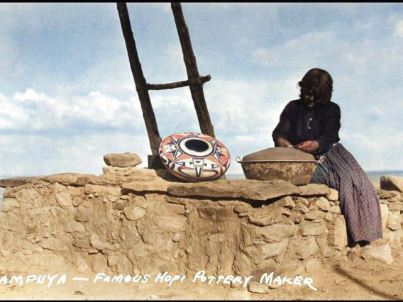 a Hopi woman sits on a rock wall forming a large, wide-shouldered jar using a large metal basin as a base