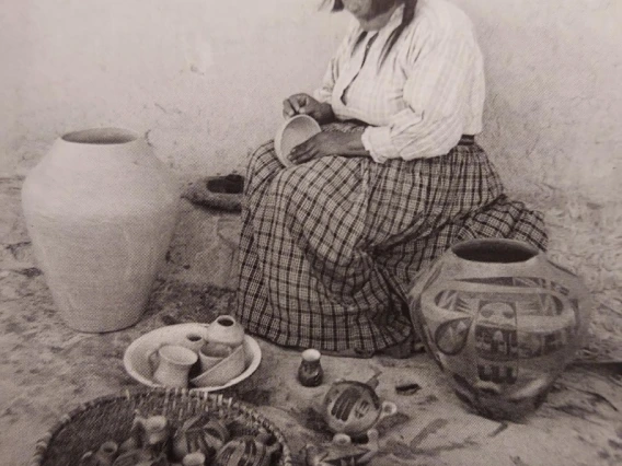 a Hopi woman is sitting outside a dwelling, on an adobe ledge, making a small bowl. She is flanked by two large ollas on either side.