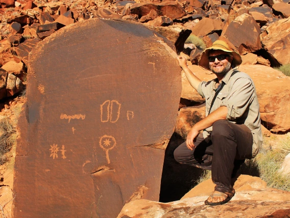 a man wearing a hat is kneeling next to a tall stone slab that shows petryglyphs on it