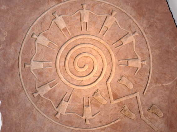 This image is of a sandstone-carved symbol illustrating the paths of life concept, with hand-holding figures standing in a circle above a spiraliform maze. The piece is by Hopi artist Gerald Dawavendewa.
