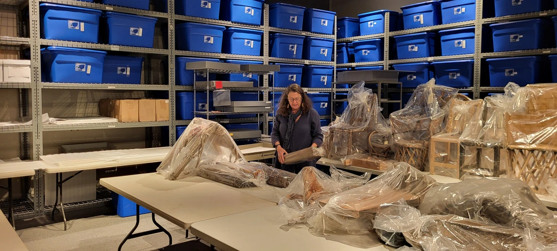 A woman is placing items on a table. Blue tubs on shelves are in the background.