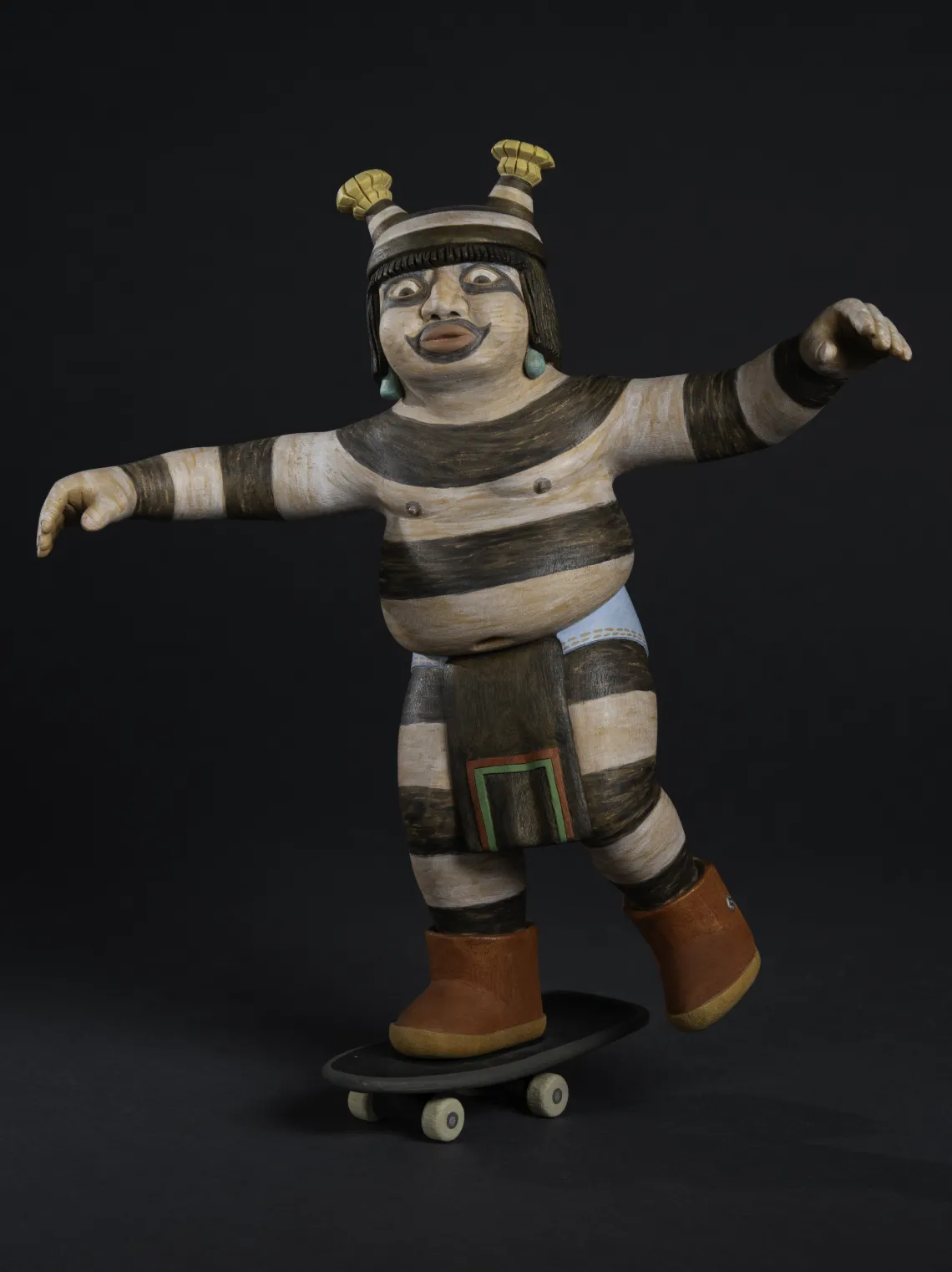 a Hopi clown with white body paint and horizontal black stipes rides a skateboard with his arms out for balance