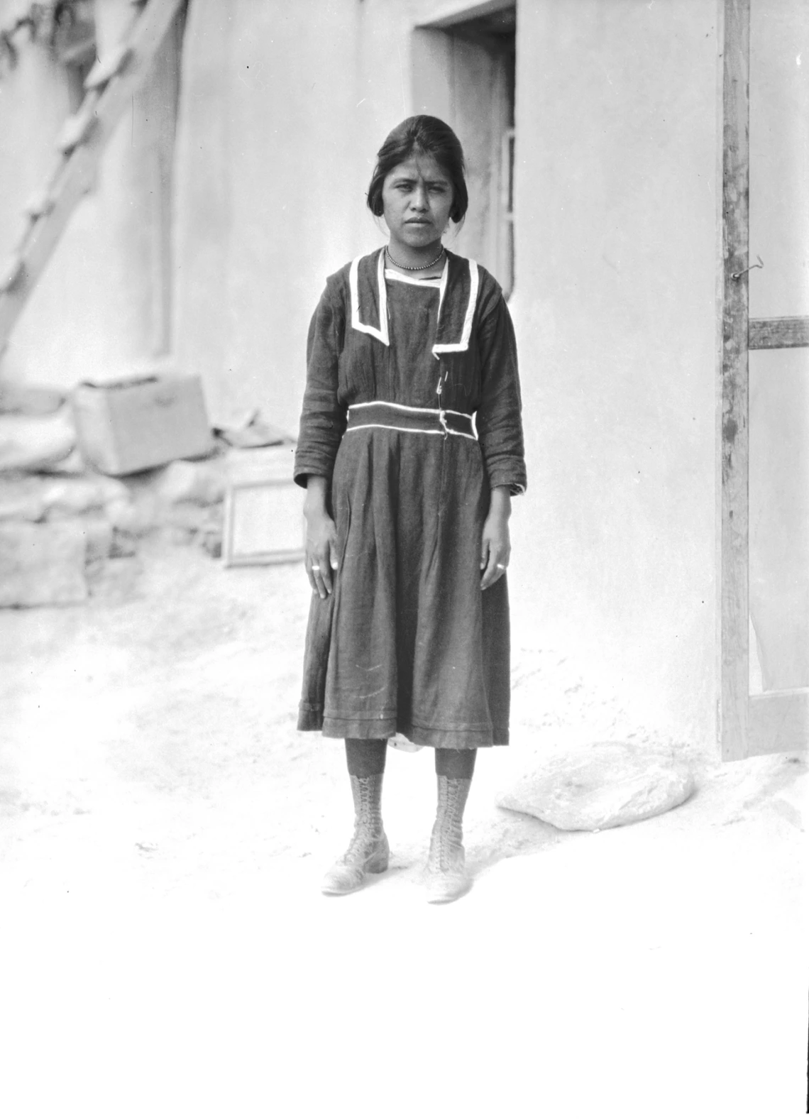 a young Hopi woman with short hair is standing outside a house and looking into the camera. She is wearing a dark, long-sleeved dress. Her button-up boots go up to her calves.