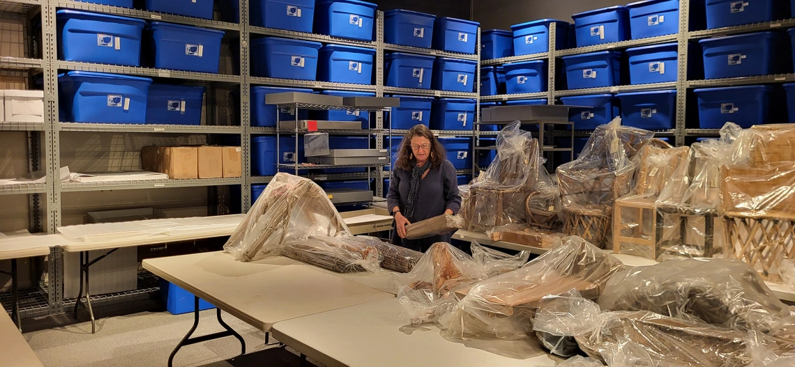 A woman is placing items on a table. Blue tubs on shelves are in the background.