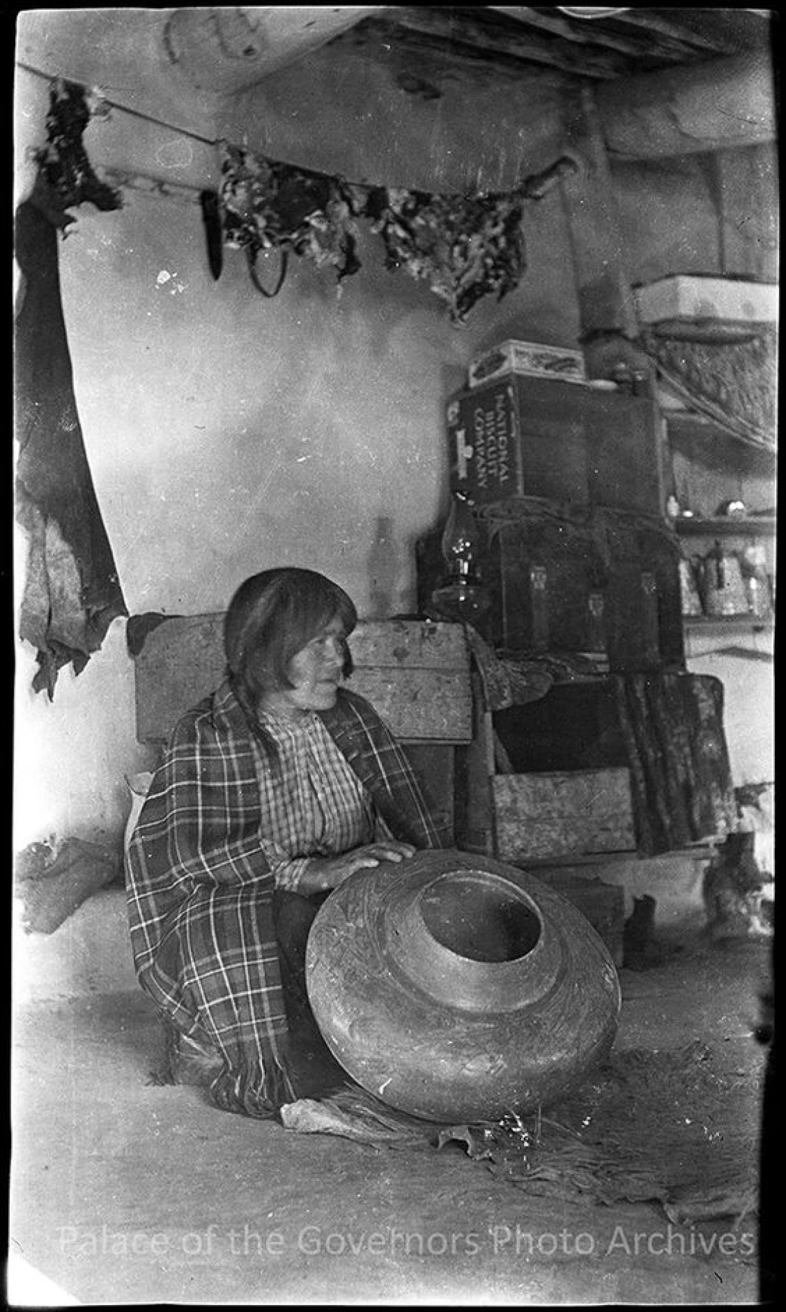 A Hopi woman with a shawl around her shoulders sits on the floor of a dwelling or store holding a large ceramic olla in front of her