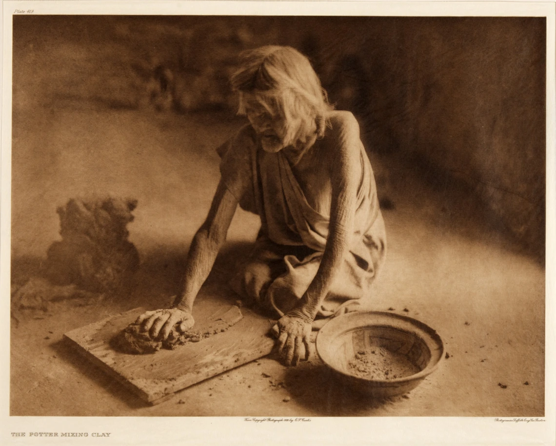 an elderly Hopi woman kneels on the ground and is kneading clay with her hands. To her side is a bows with an everted rim.