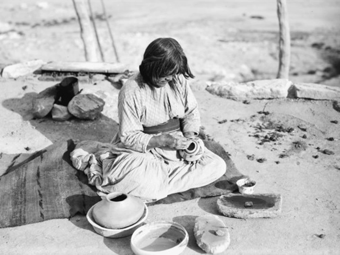 A Hopi woman sits on the ground in the beginning stages of making a pot.