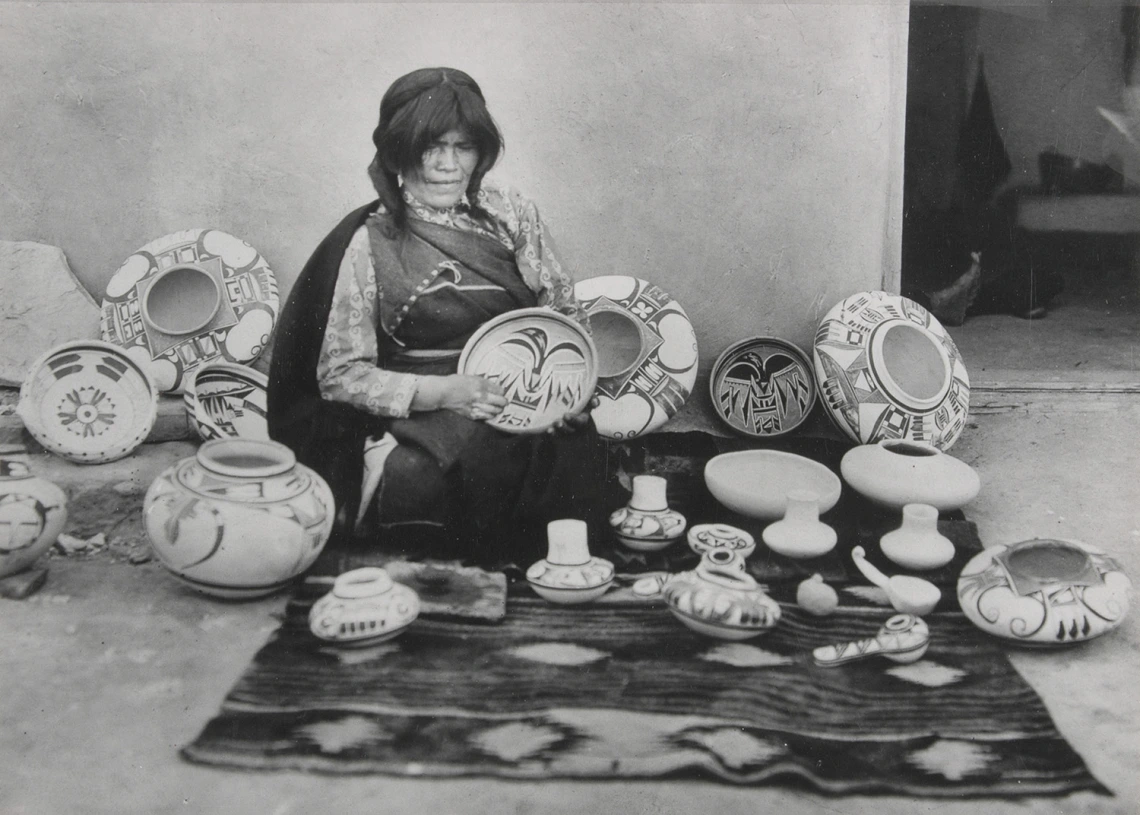 a Hopi woman sits on the ground with a blanket spread before her and a great number of pots she has made surround her