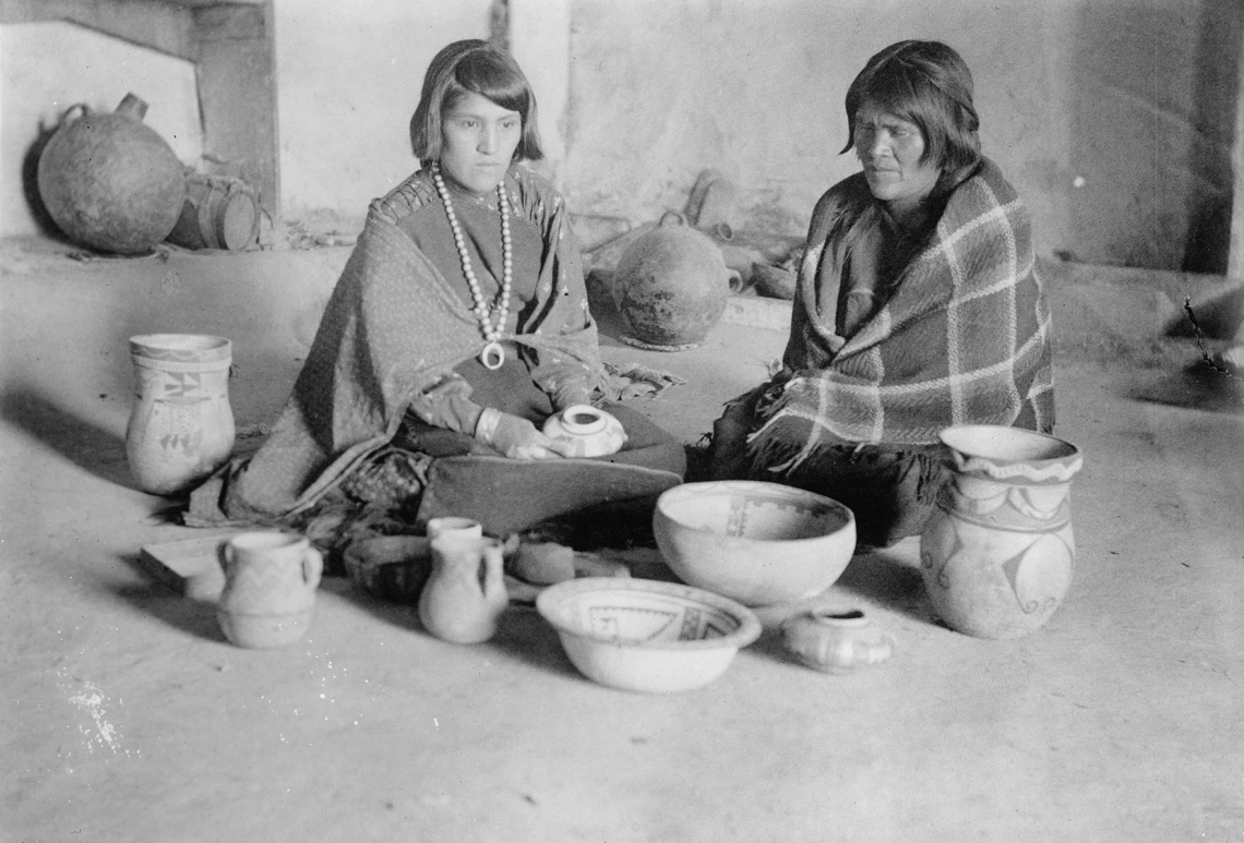 a young Hopi woman with short hair shits on the ground looking at pottery she and her mother, to her left, have made together