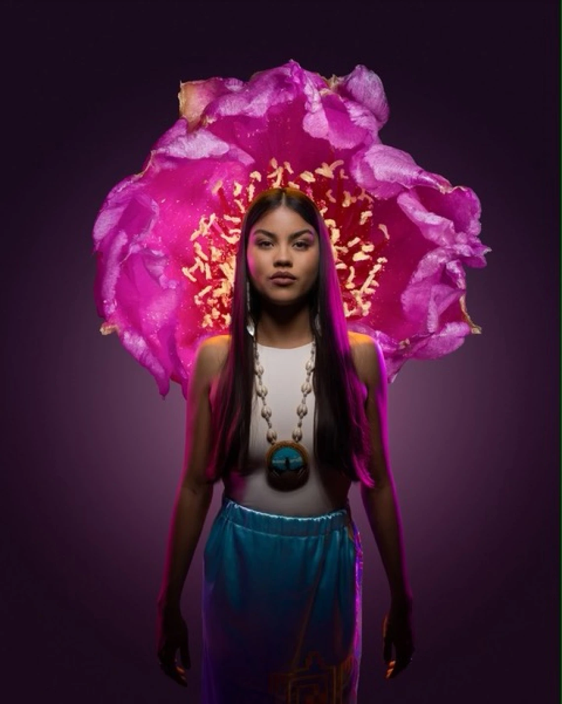 a young woman with long dark hair stands in front of a large bright pink flower