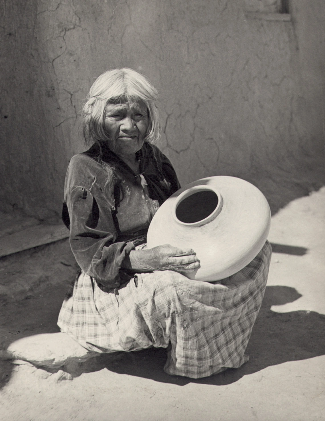 An elderly Hopi woman sits on a low stoop, holding a ceramic jar she is in the process of making.