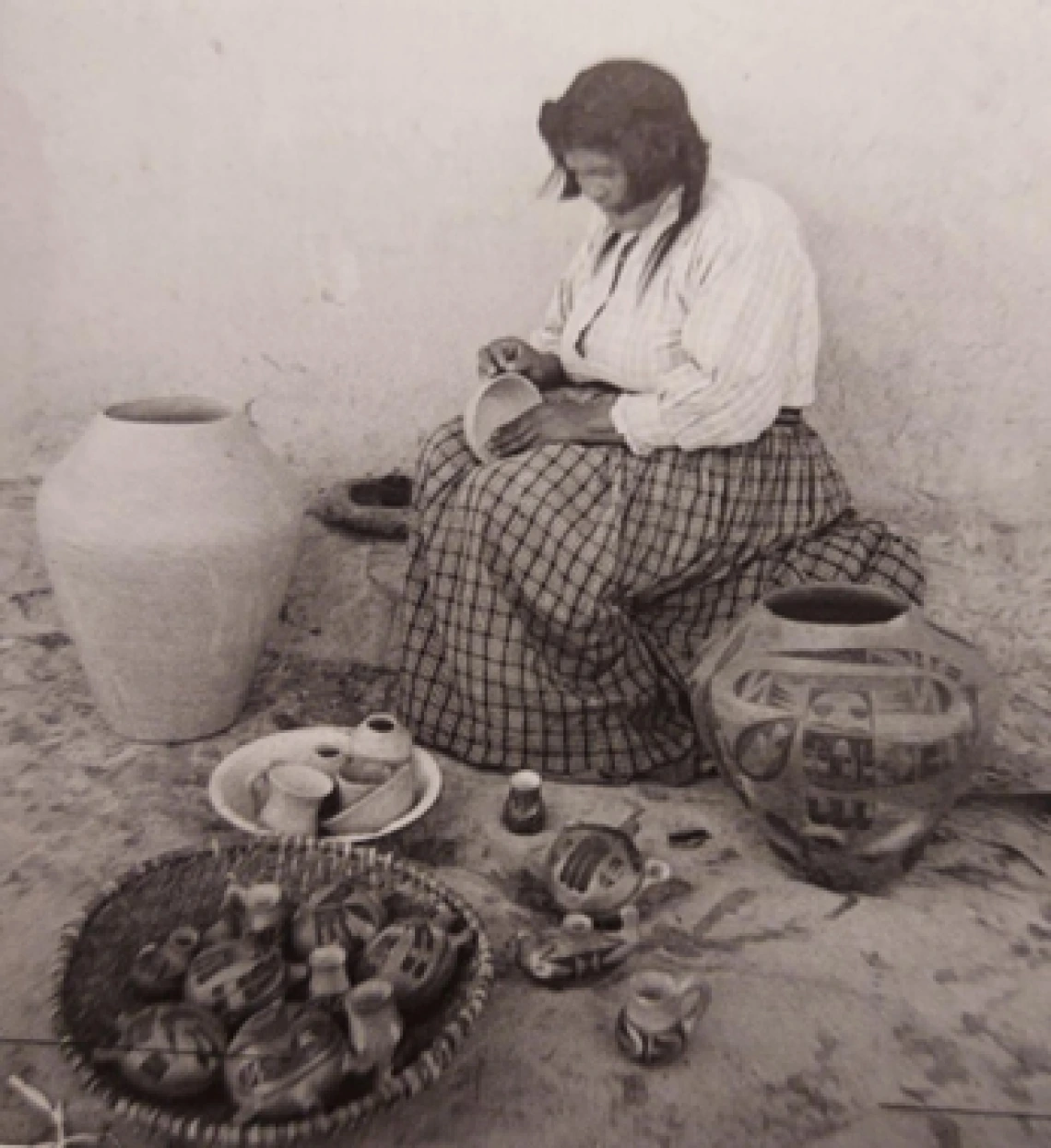 a Hopi woman is sitting outside a dwelling, on an adobe ledge, making a small bowl. She is flanked by two large ollas on either side.