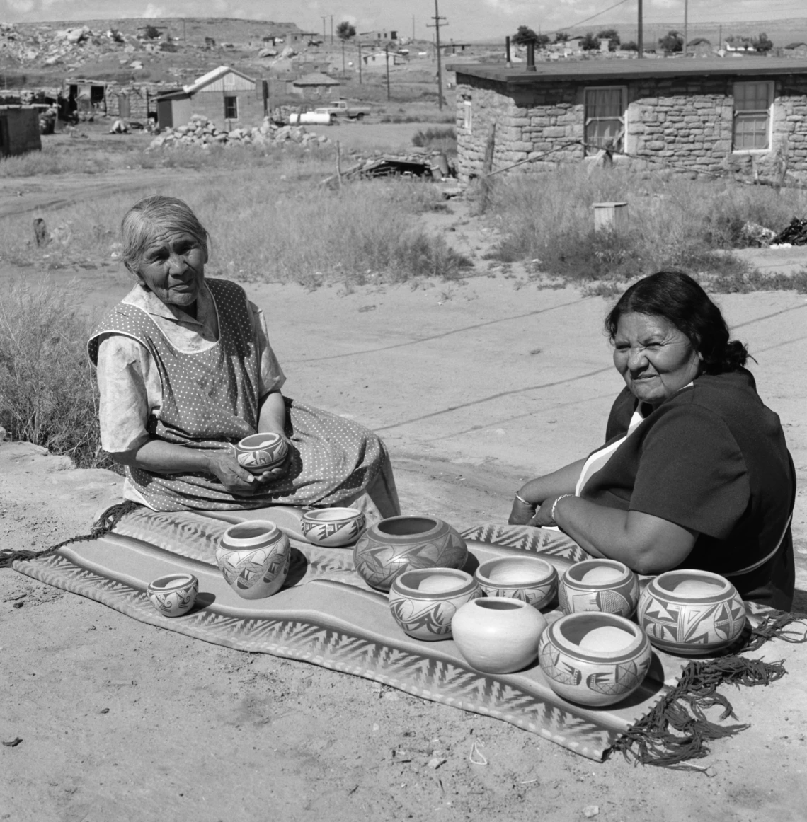 two Hopi women are sitting outside in front of a ledge on which are ten ceramic vessels spread out on a blanket