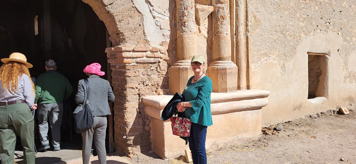 A woman wearing a green tunic top and wearing a baseball cap smiles at the camera at Tumacacori National Historical Park.