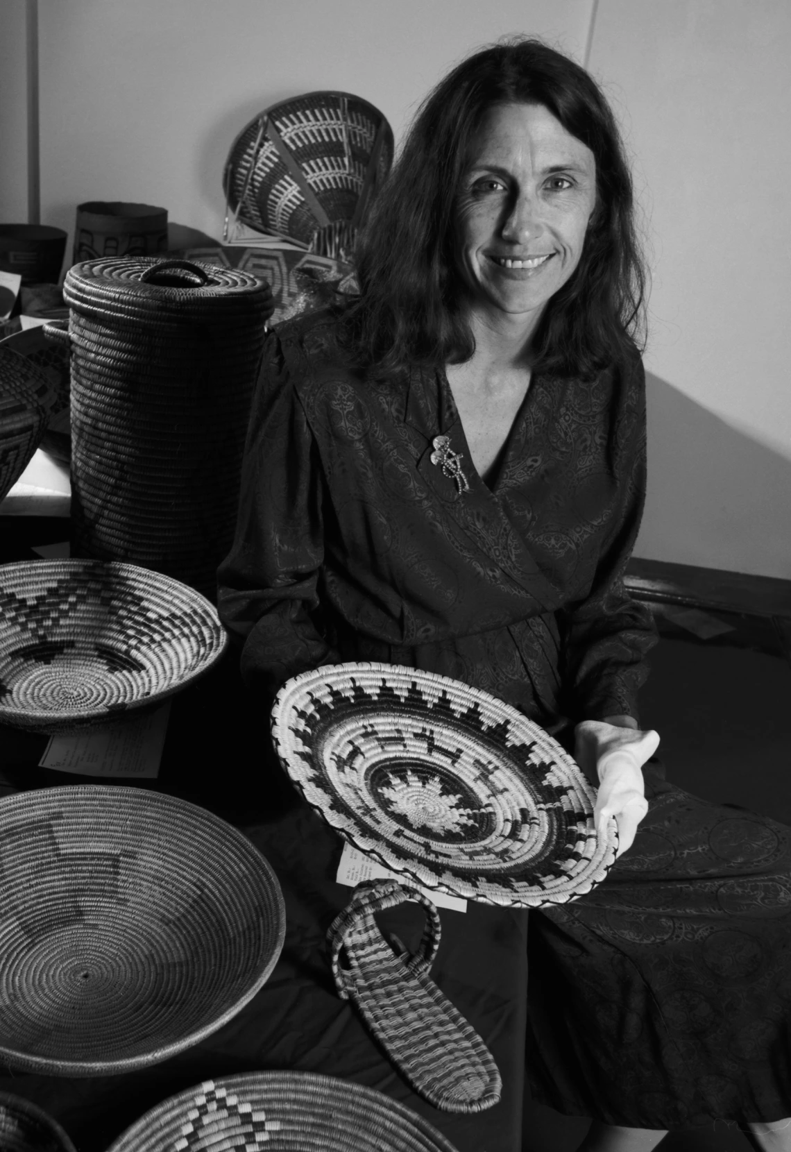 A woman sits holding a basket, surrounded by other baskets.