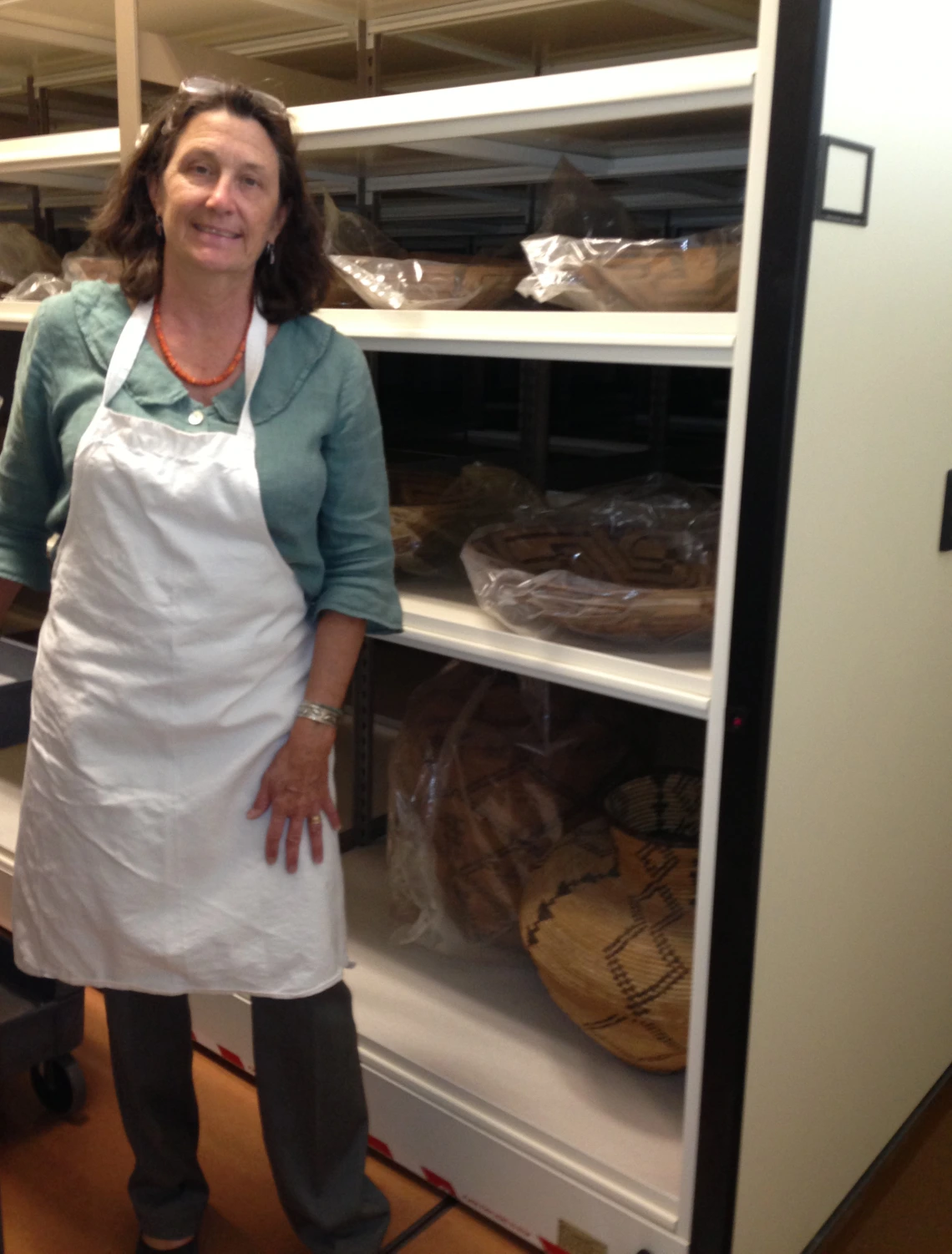 A woman wearing a white apron over a green shirt peeks out of an aisle in a basketry vault.