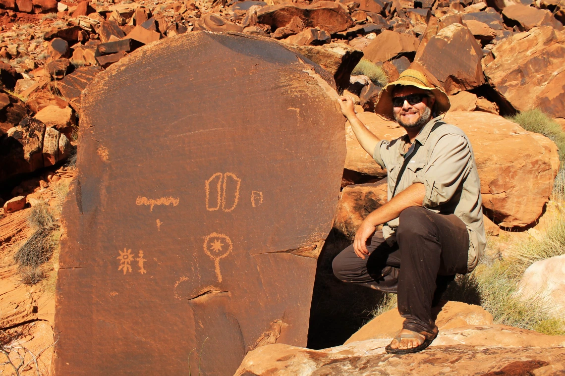 a man wearing a hat is kneeling next to a tall stone slab that shows petryglyphs on it