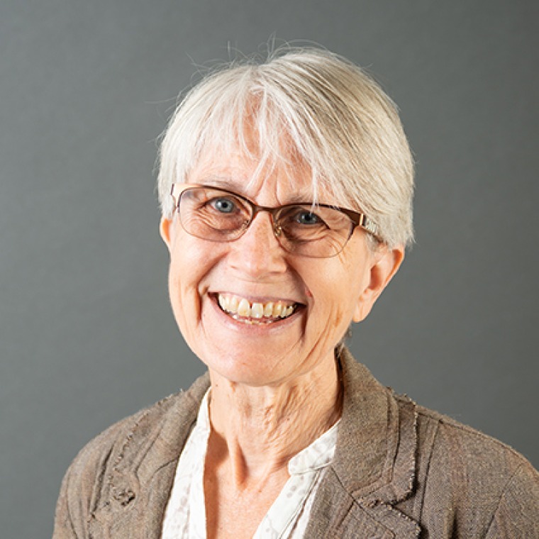 Woman smiling into the camera wearing glasses. Her white hair is very short. She is wearing a khaki-colored jacket over a white blouse.