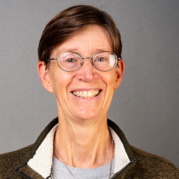 Woman smiling into the camera wearing glasses. Her brown hair is very short. She is wearing a brown-colored jacket over a grey top. 