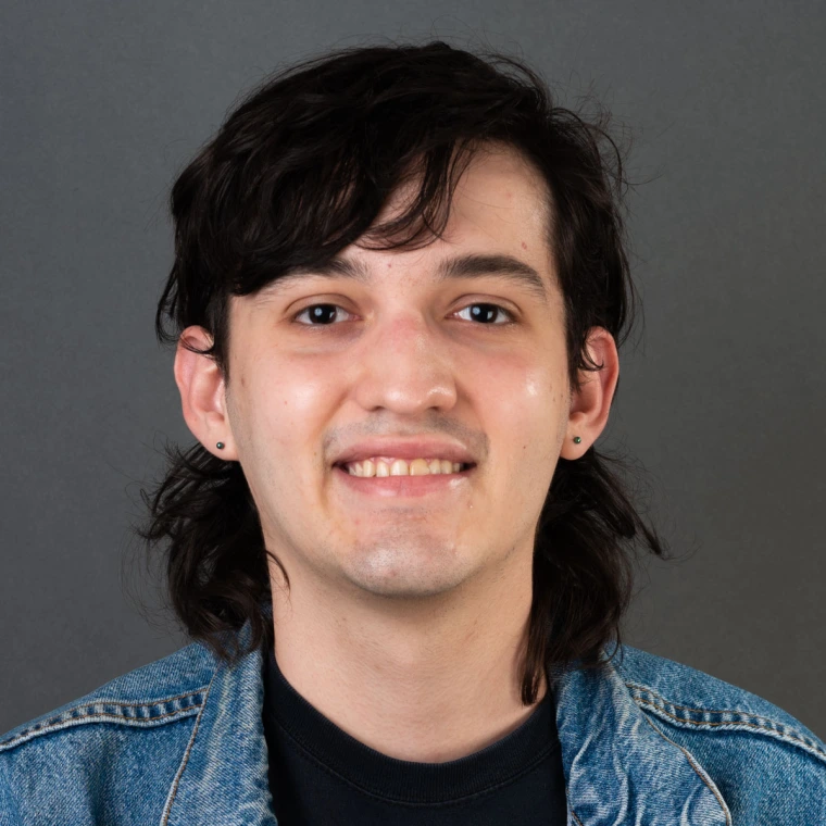 Young man with dark, shoulder-length hair smiles into the camera wearing a denim jacket over a dark blue shirt.