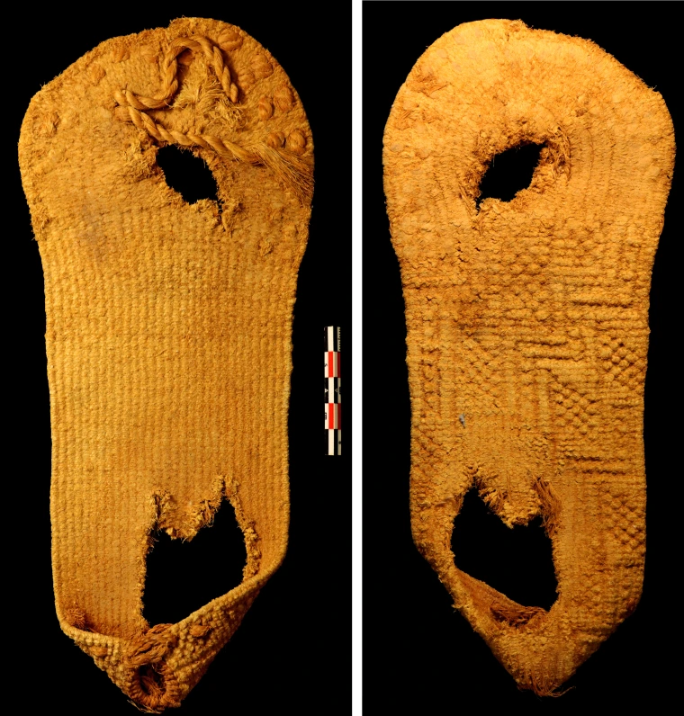The sandal, like all the sandals in this exhibit, is made out of yucca fiber which is an ochre shade of orange. It has a round toe and a puckered heel shape. The tattered remains of a toe strap are visible. It has a large hole under the heel and a smaller hole under the ball of the foot.