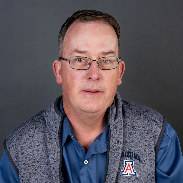 a man with very short brown hair and glasses is wearing a heather-grey UA sweater vest over a light blue button-down shirt