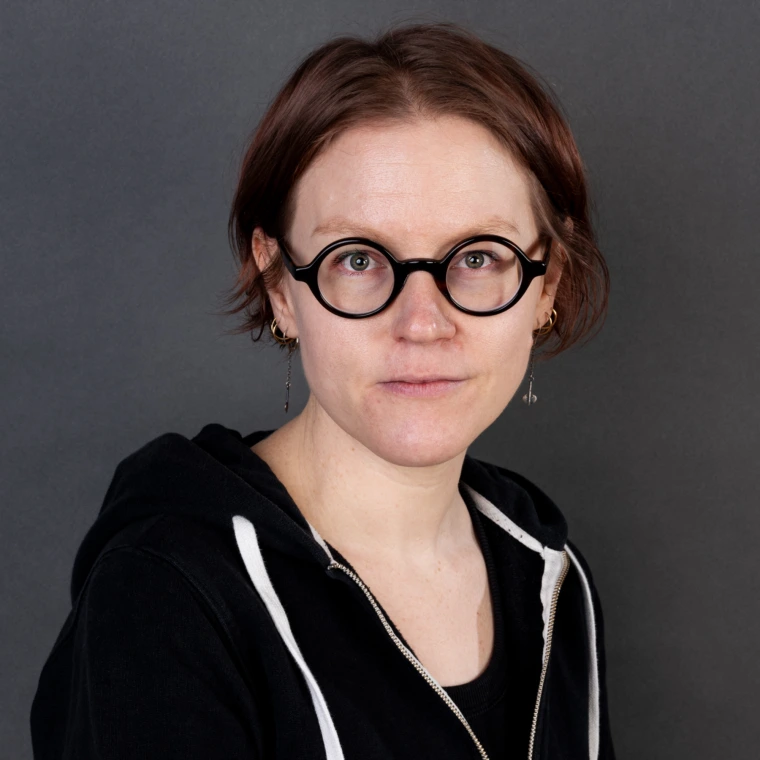 a woman with ear-length brown-red hair and round black-framed glasses is wearing a black zip-up hooded sweater with white trim