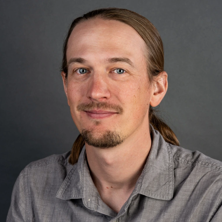 a man with a ponytail, moustache, and goatee is waring a gray button down shirt