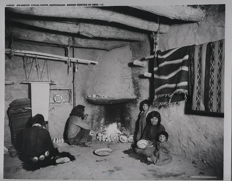 A Hopi family sit on a dirt floor. A male is weaving cotton on the left. A man tends a fireplace in the center. To the right, a woman holds a ceramic bowl. She has two children by her side.