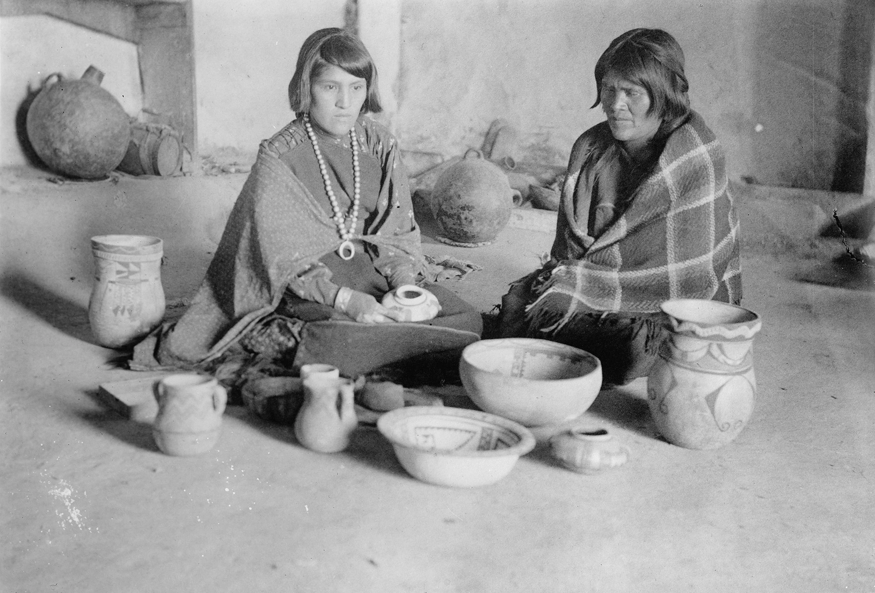 a young Hopi woman with short hair shits on the ground looking at pottery she and her mother, to her left, have made together