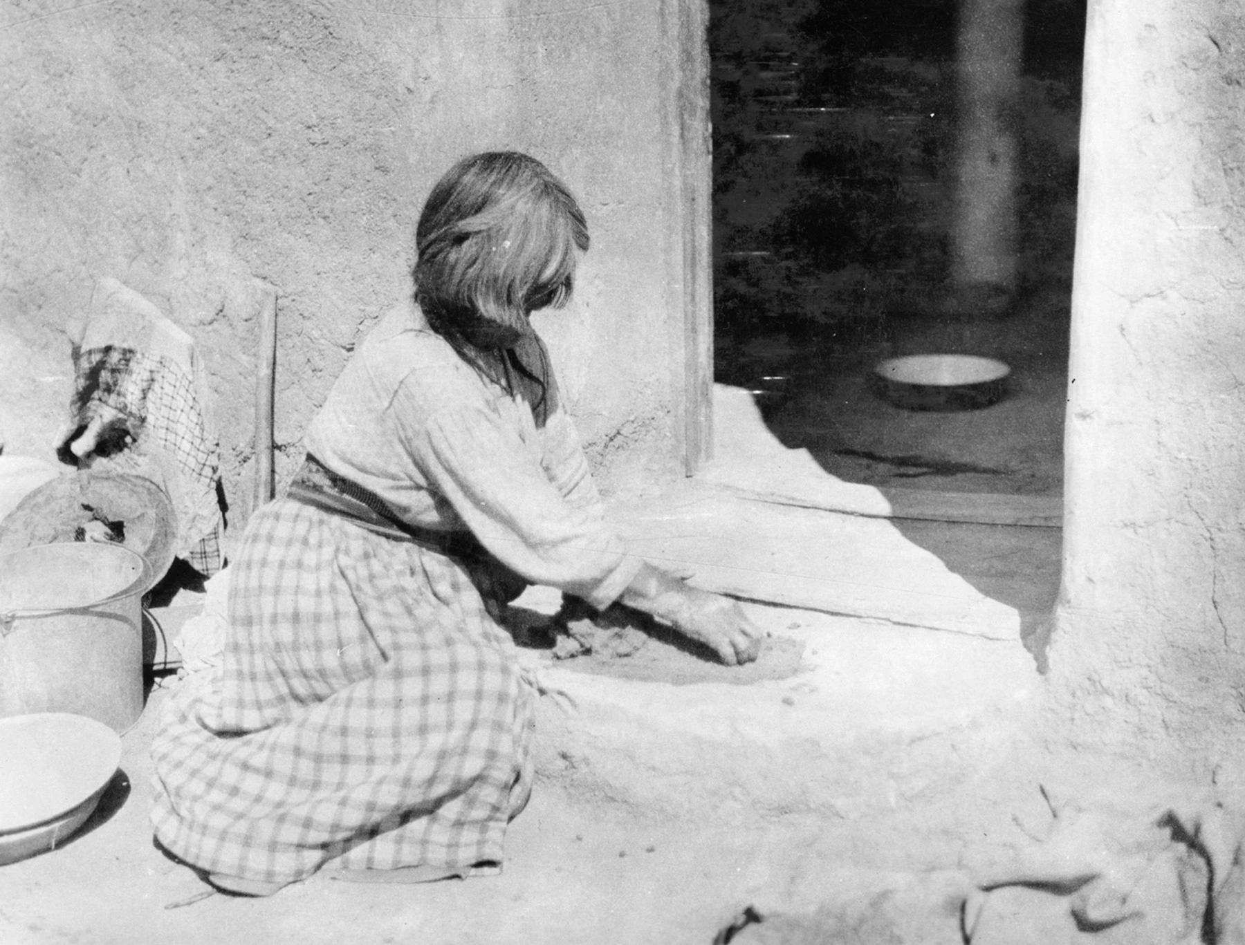 a woman is sitting on the ground, outside the doorway of a dwelling, kneading clay in the sun