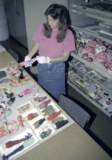 A woman stands amid an array of folk art in front of her on a table and in back of her in cabinet drawers.