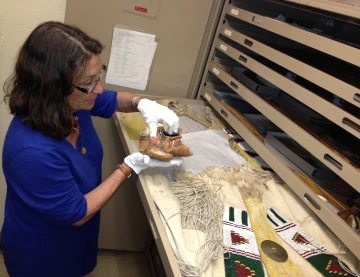 A woman stands in front of a drawer full of beaded artifacts. She is holding a pair of moccasins.