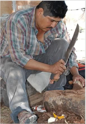 A man uses a machete to fashion wood into the shape of a mask
