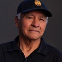 Man wearing a dark blue polo shirt, his silver sideburns can be seen under a dark blue baseball cap.