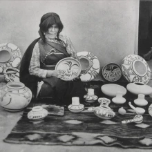 a Hopi woman sits on the ground with a blanket spread before her and a great number of pots she has made surround her